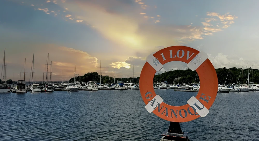 A scenic view of a marina in Gananoque at sunset, with sailboats docked under a sky of dramatic orange and grey clouds. In the foreground, an orange lifebuoy on a post is inscribed with the message "# I LOVE GANANOQUE."